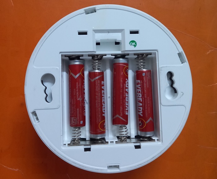 Insert batteries inside a wireless smoke detector with polarities visible. 